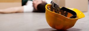 A helmet on the floor with a worker lying unconscious in the background.