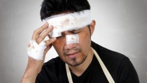 A man with a bandage around his head, nose, and hand holding his head in pain. 