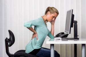 A woman sat at a computer desk holding her back.