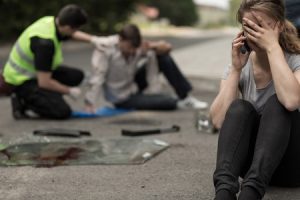A person sat down and making a phone call. Behind them, a pedestrian hit by a car is seated and being attended to by a person in a hi-viz jacket.