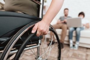 A close-up image of a person sat in a wheelchair with two people sat on a sofa in the background.