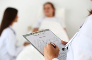 A doctor writing on a clipboard with a patient in the background.