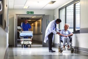 A hospital corridor with a doctor speaking to a patient in a wheelchair to the right and a nurse pushing a bed in the middle distance.