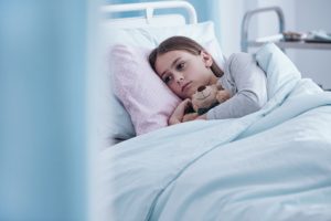A girl in a hospital bed holding a teddy bear under her arm.