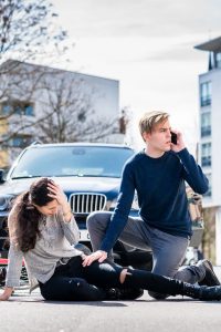 A man phoning for help with crouching next to a woman who has been hit by a car.