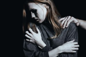 Dark image of a woman holding herself and crying while a hand comforts her from behind. 