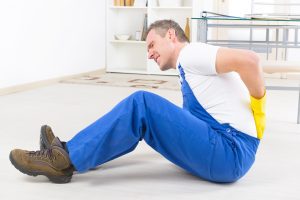 A man in blue dungarees sat on the floor holding his back.