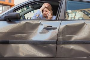 A woman looking distressed on the phone inside a car that has a dent in the door.