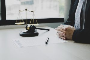 A Solicitor Sits With Folded Hands At A Desk with A Gavel And Scales. 