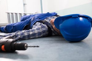 A man with a checked shirt, blue overalls and a blue helmet lying on his back on the floor, with a screwdriver next to him.