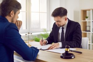 A logistics accident compensation claim solicitor, wearing a dark suit and tie, sits at a desk and writes in a legal pad. Their client sits across from them and is wearing a blue suit.