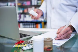A pharmacist stands at a desk holding a bottle of pills and filling out a prescription.