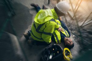 A man in construction gear with his helmet fallen off lying on his back, and another construction worker over him. 