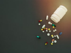 A small white plastic tub knocked over with a bunch of splled pills, all a variety of shapes and colours, with a grey background. 