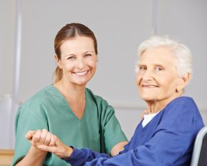 An elderly woman smiling sat down holding hands with a young female carer also smiling sat down, wearing a green uniform. 