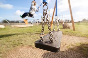 An empty swing set in the foreground with a child swinging on a swing in the background.