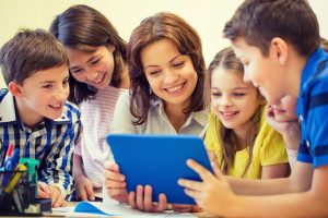 A teacher is surrounded by students as they read a tablet