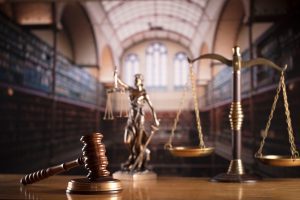 A close up of a gavel, statue, and scales with a court blurred out in the background.