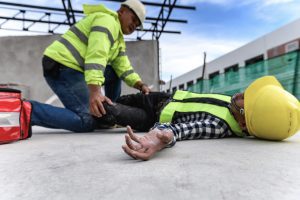 A construction employee lies on the ground while their colleague tries to help them after an accident happened. The employee may be able to claim compensation for their medical treatment