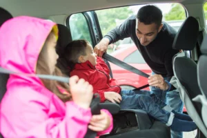 A father securing his young son in a booster seat while his daughter buckles her own seat belt