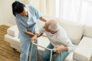A caregiver helps a patient move from a white sofa.